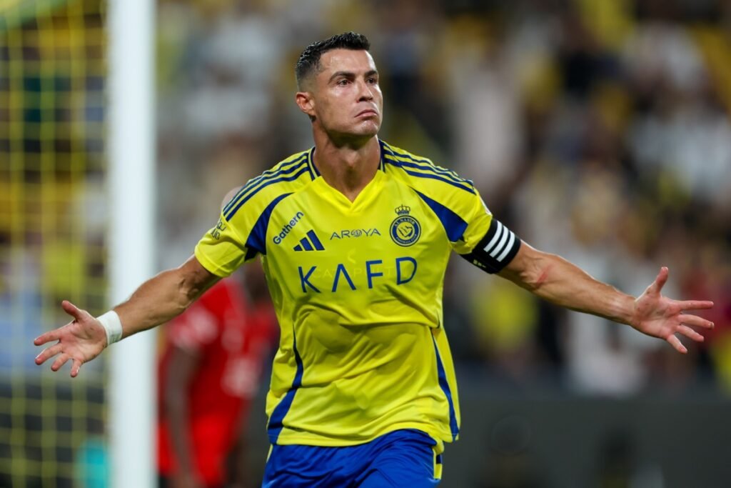 Jogador de futebol comemorando gol com os braços abertos, vestindo uniforme amarelo e azul em campo de futebol, expressão intensa.