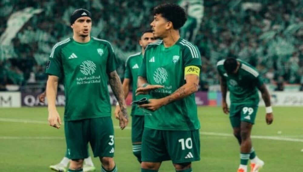 Jogadores de futebol em campo usando uniformes verdes, um deles usando braçadeira de capitão, discutem estratégia durante o jogo.