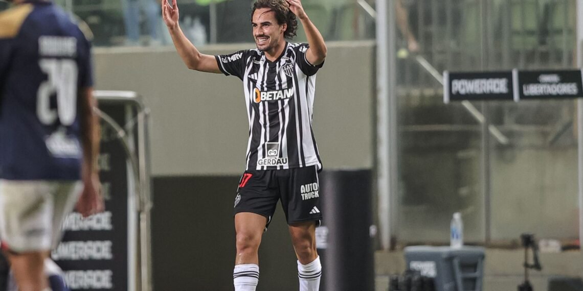 Jogador comemorando gol em campo, vestindo uniforme listrado preto e branco, torcida ao fundo. Partida de futebol.