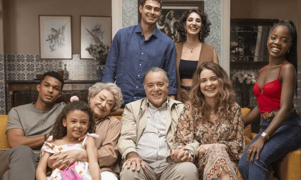 Família feliz reunida no sofá, composta por várias gerações, sorrindo em ambiente acolhedor e decorado com quadros.