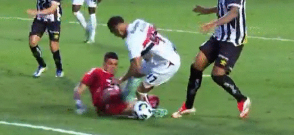 Jogador de futebol em lance disputado, com goleiro de uniforme vermelho tentando agarrar a bola durante um jogo competitivo.