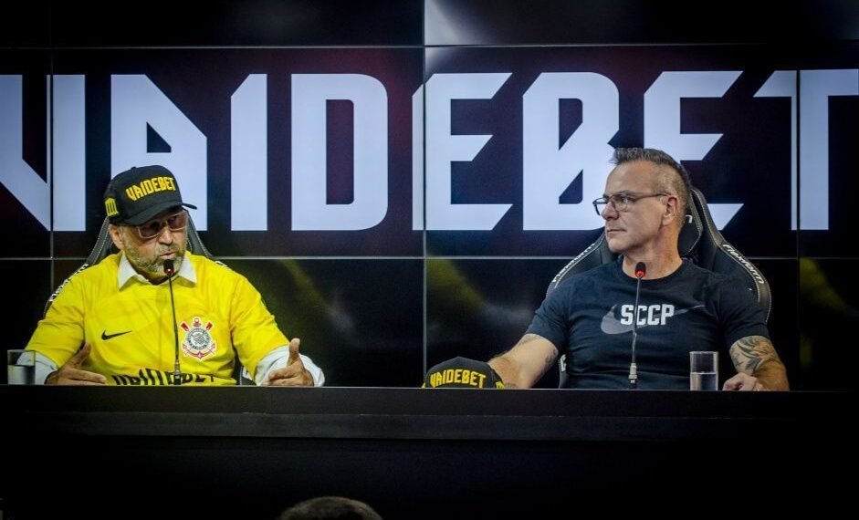 Dois homens em mesa de coletiva de imprensa do Corinthians, usando camisetas e bonés de patrocinador, em frente a painel.