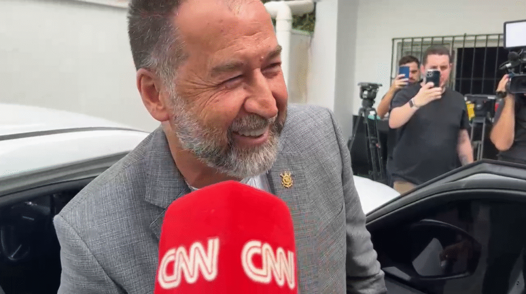 Homem sorridente em entrevista, próximo a carro. Equipe de filmagem e fotógrafos ao fundo registrando o momento.