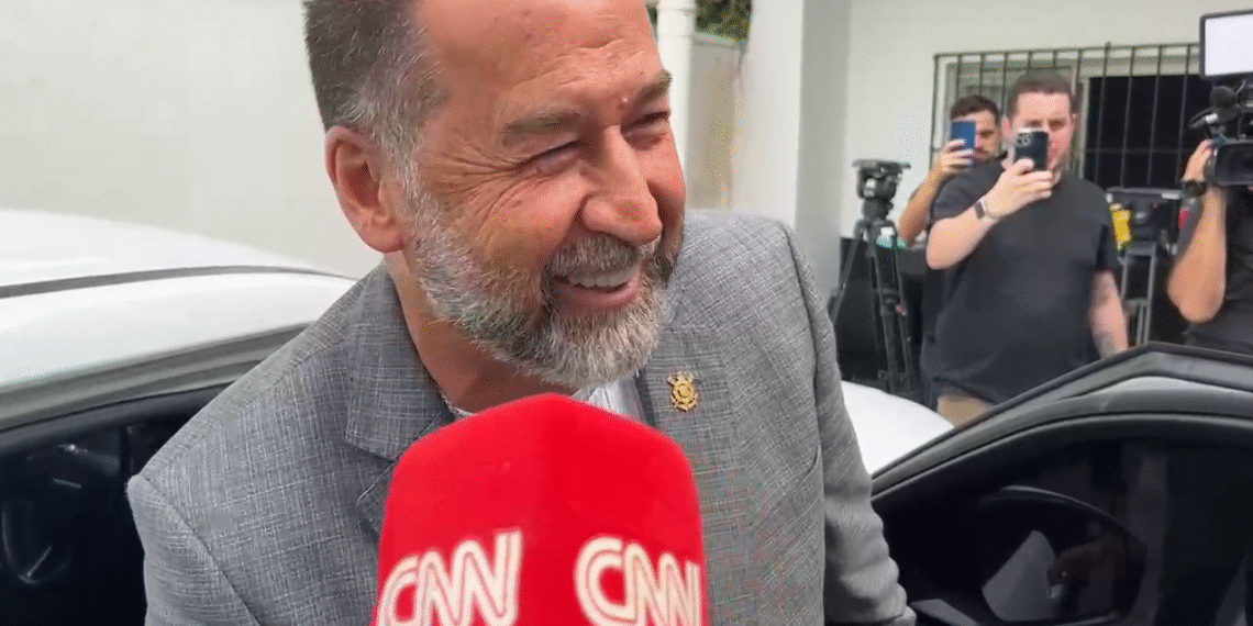 Homem sorridente em entrevista, próximo a carro. Equipe de filmagem e fotógrafos ao fundo registrando o momento.