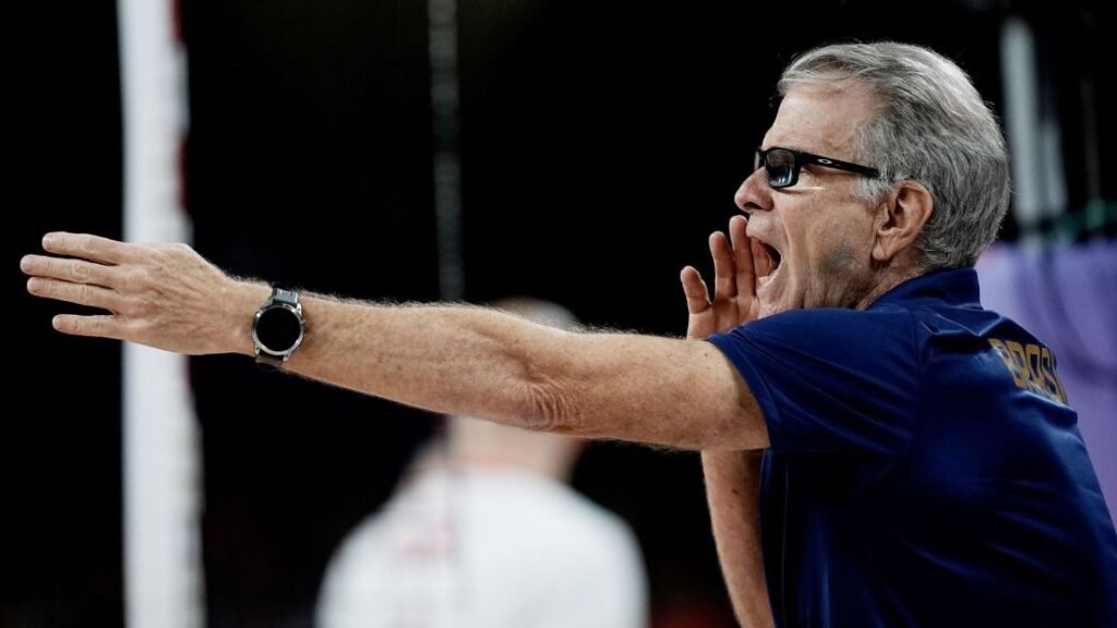 Técnico de vôlei grita instruções durante jogo, usando camisa azul com logotipo atrás, gesticulando com um braço estendido.