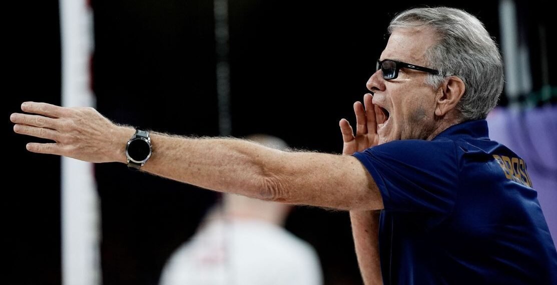 Técnico de vôlei grita instruções durante jogo, usando camisa azul com logotipo atrás, gesticulando com um braço estendido.