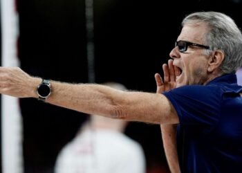 Bernardinho reúne craques para a Seleção Brasileira de Vôlei 32 Técnico de vôlei grita instruções durante jogo, usando camisa azul com logotipo atrás, gesticulando com um braço estendido.
