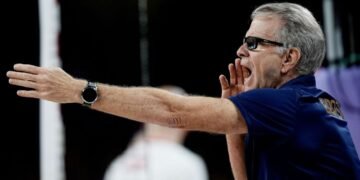 Técnico de vôlei grita instruções durante jogo, usando camisa azul com logotipo atrás, gesticulando com um braço estendido.