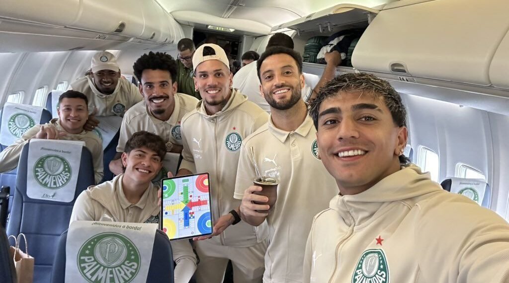 Jogadores do Palmeiras posam sorridentes em um avião, interagindo com um tablet e uniformizados, em clima de viagem.