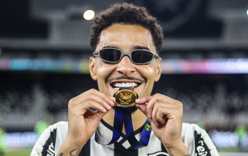 Pessoa sorrindo, usando óculos escuros, exibe medalha de ouro com fita azul em estádio, celebrando conquista esportiva.