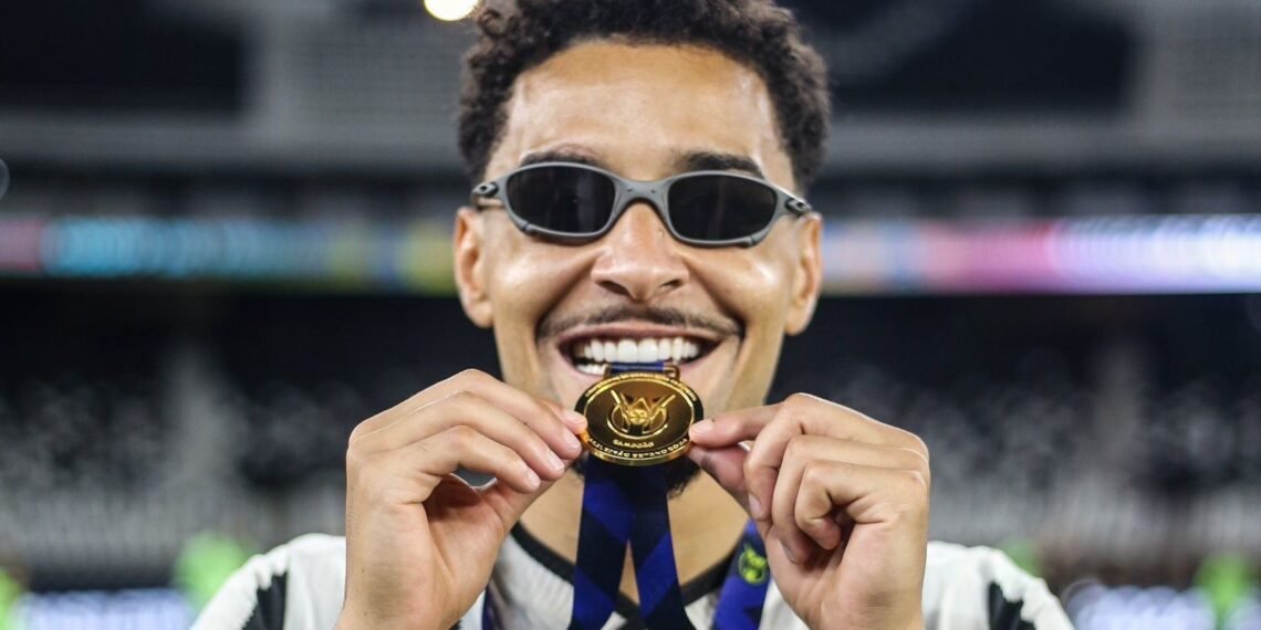 Pessoa sorrindo, usando óculos escuros, exibe medalha de ouro com fita azul em estádio, celebrando conquista esportiva.