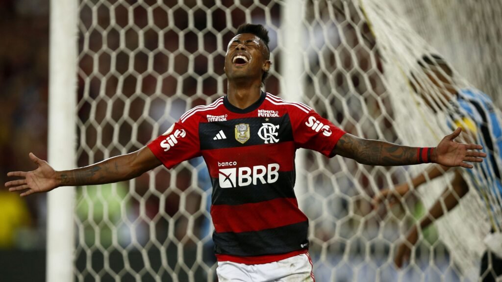 Jogador de futebol do Flamengo comemora gol com expressão de alegria em frente ao gol, braços abertos, em campo de futebol.
