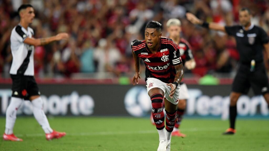Jogador do Flamengo em ação durante partida, vestindo uniforme com listras vermelhas e pretas, corre pelo campo atento ao jogo.