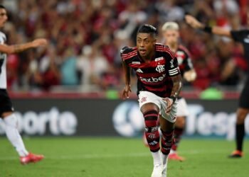 Bruno Henrique comenta sobre apuração de apostas suspeitas 39 Jogador do Flamengo em ação durante partida, vestindo uniforme com listras vermelhas e pretas, corre pelo campo atento ao jogo.