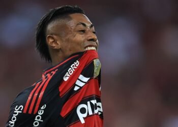 Jogador de futebol do Flamengo, em close, com expressão intensa, segura a camisa vermelha e preta durante o jogo.
