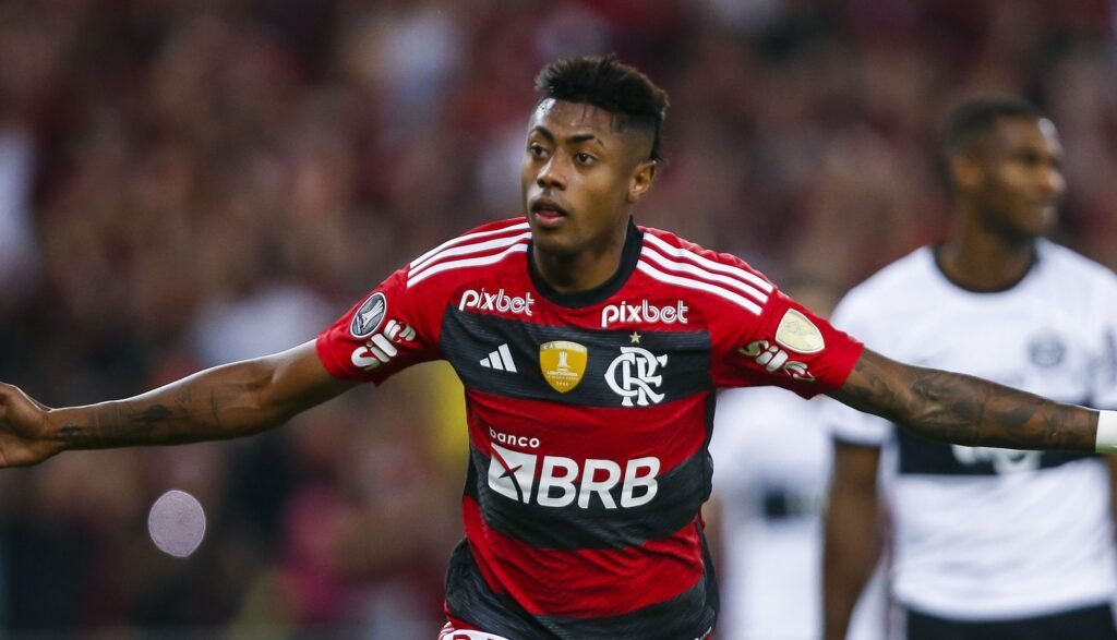 Jogador de futebol do Flamengo comemora gol em campo, vestindo uniforme vermelho e preto, torcedores ao fundo.