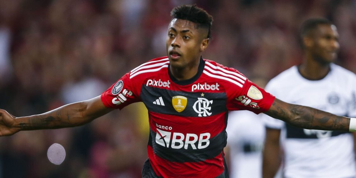 Jogador de futebol do Flamengo comemora gol em campo, vestindo uniforme vermelho e preto, torcedores ao fundo.
