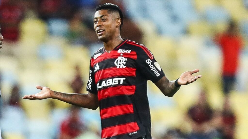 Jogador do Flamengo em campo, com expressão de dúvida, vestindo uniforme rubro-negro, em estádio iluminado.