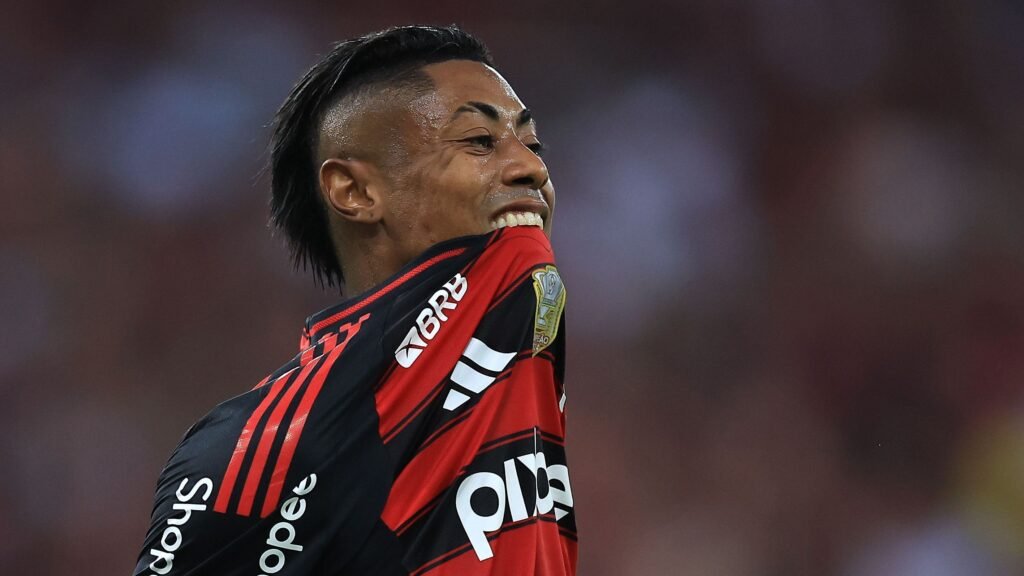 Jogador de futebol em campo morde a própria camisa em um gesto intenso, vestido com uniforme vermelho e preto.