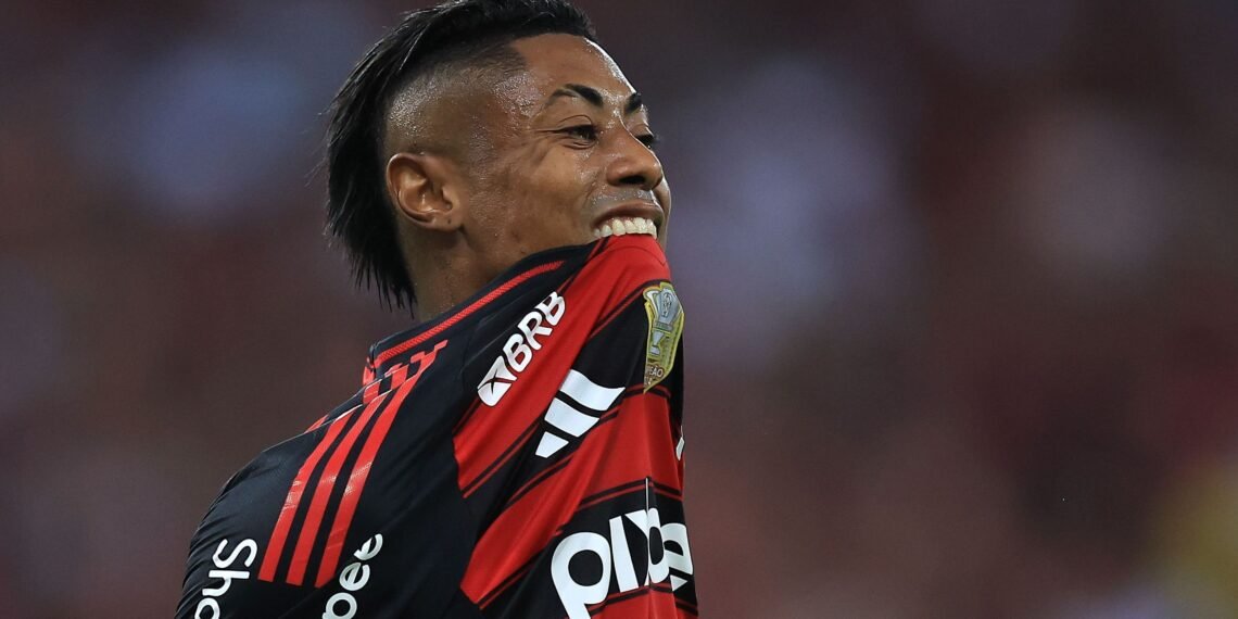 Jogador de futebol em campo morde a própria camisa em um gesto intenso, vestido com uniforme vermelho e preto.