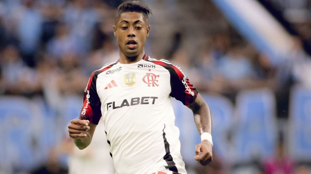 Jogador do Flamengo em ação durante partida de futebol, vestindo uniforme branco, com expressão concentrada e fundo desfocado.
