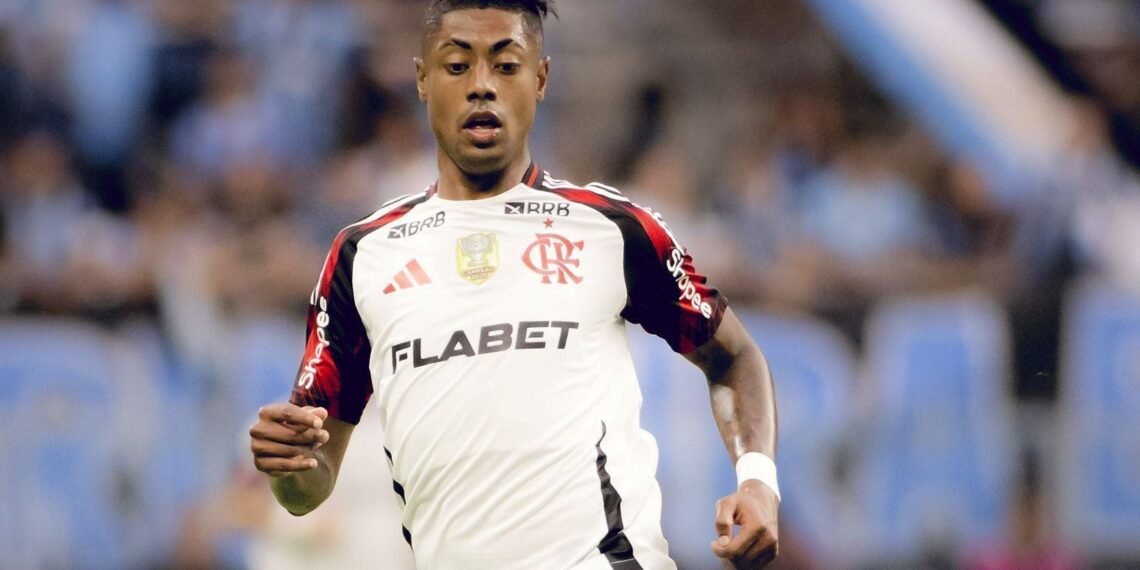 Jogador do Flamengo em ação durante partida de futebol, vestindo uniforme branco, com expressão concentrada e fundo desfocado.