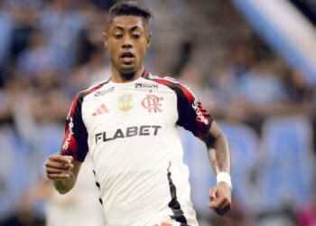 Jogador do Flamengo em ação durante partida de futebol, vestindo uniforme branco, com expressão concentrada e fundo desfocado.