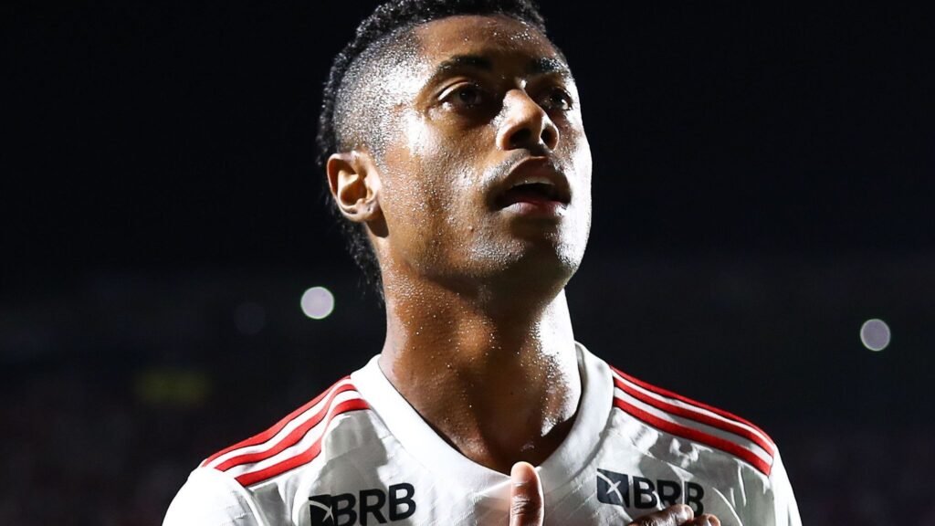 Jogador de futebol em destaque, com expressão concentrada, veste uniforme branco com detalhes vermelhos em partida noturna.