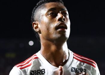 Jogador de futebol em destaque, com expressão concentrada, veste uniforme branco com detalhes vermelhos em partida noturna.