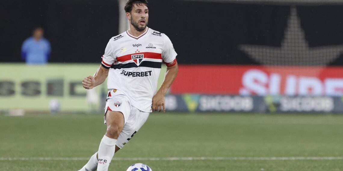 Jogador de futebol do São Paulo conduz a bola em campo durante partida, vestindo uniforme branco com detalhes vermelhos e pretos.