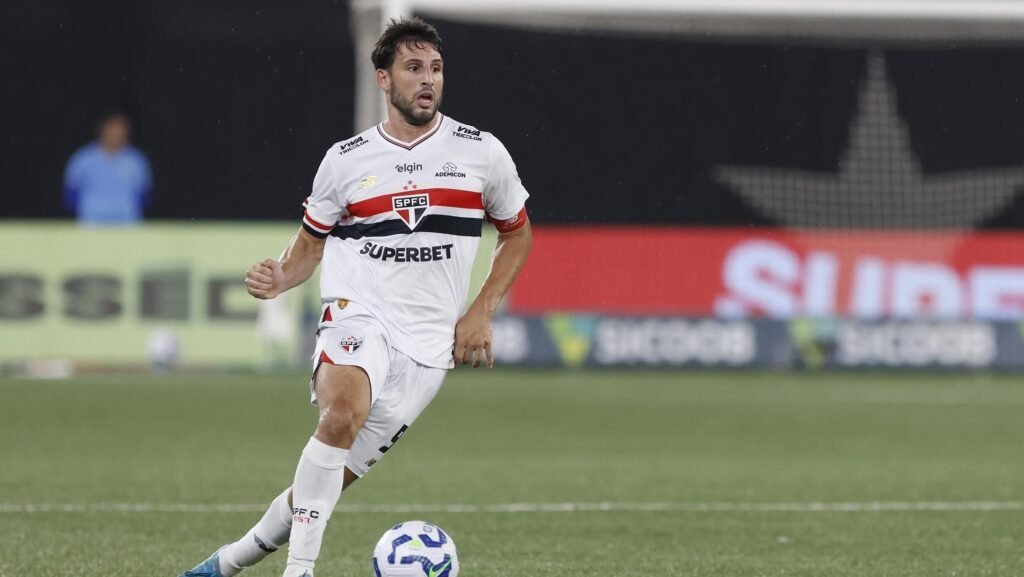 Jogador de futebol do São Paulo correndo, com bola nos pés, em campo durante partida, usando uniforme branco do time.