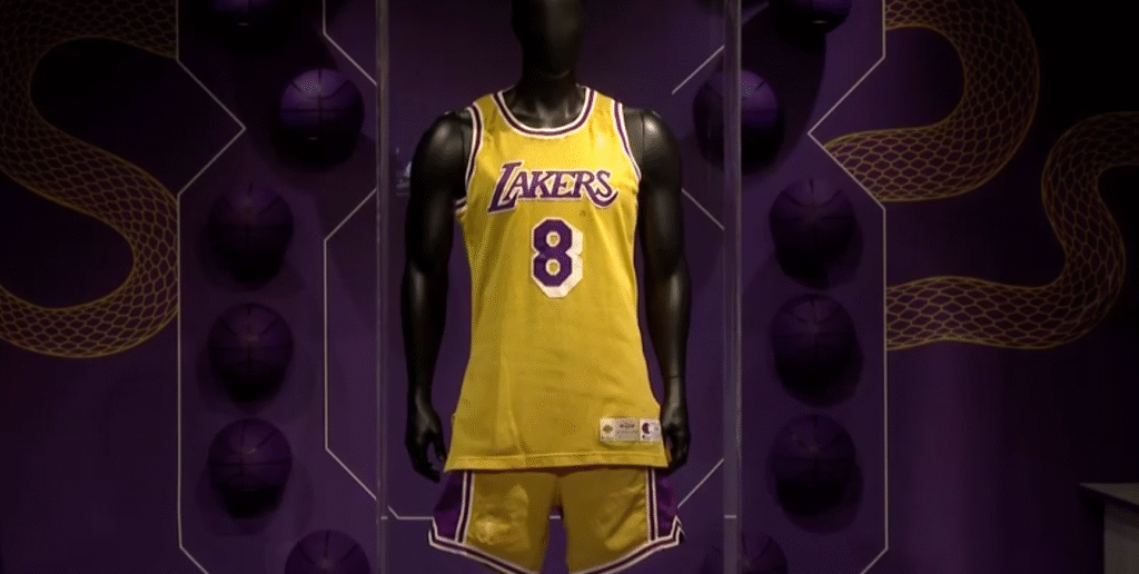 Uniforme amarelo dos Lakers com número 8 em manequim, exibido em cenário temático de basquete com bolas roxas decorativas.