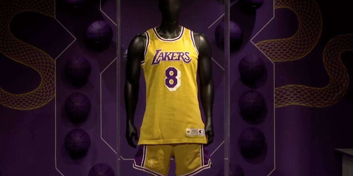 Uniforme amarelo dos Lakers com número 8 em manequim, exibido em cenário temático de basquete com bolas roxas decorativas.