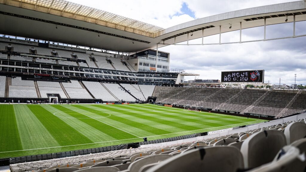 Estádio de futebol moderno vazio, com grama bem cuidada e arquibancadas brancas, sob um céu nublado.
