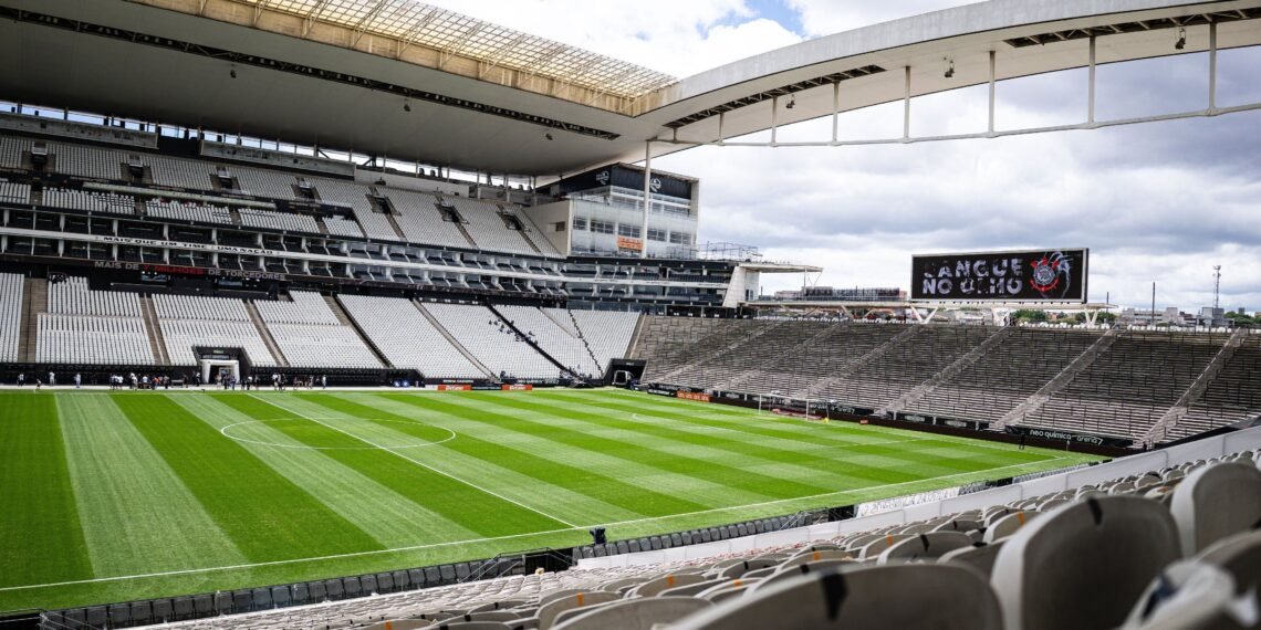 Estádio de futebol moderno vazio, com grama bem cuidada e arquibancadas brancas, sob um céu nublado.