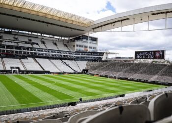 Campanha do Corinthians para quitar estádio ultrapassa R$ 10 mi 38 Estádio de futebol moderno vazio, com grama bem cuidada e arquibancadas brancas, sob um céu nublado.