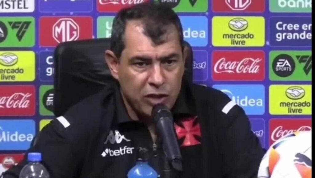 Treinador do Vasco dá entrevista coletiva após o jogo, com patrocinadores ao fundo, abordando temas esportivos.