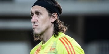 Goleiro de camisa amarela com faixas laranjas, expressão séria, faixa preta na cabeça; fundo desfocado.