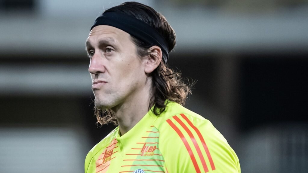 Goleiro de cabelo comprido e faixa preta veste uniforme amarelo fluorescente durante jogo, demonstrando concentração.