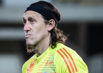 Goleiro de cabelo comprido e faixa preta veste uniforme amarelo fluorescente durante jogo, demonstrando concentração.