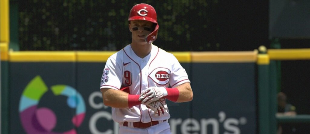 Jogador de beisebol com uniforme branco do Reds prepara-se para o jogo, usando capacete vermelho e luvas, em campo aberto.