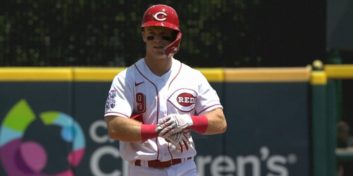 Jogador de beisebol com uniforme branco do Reds prepara-se para o jogo, usando capacete vermelho e luvas, em campo aberto.