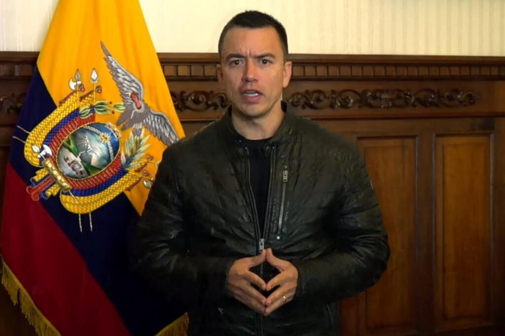 Homem em jaqueta preta fala seriamente em frente à bandeira do Equador, com madeira entalhada ao fundo.