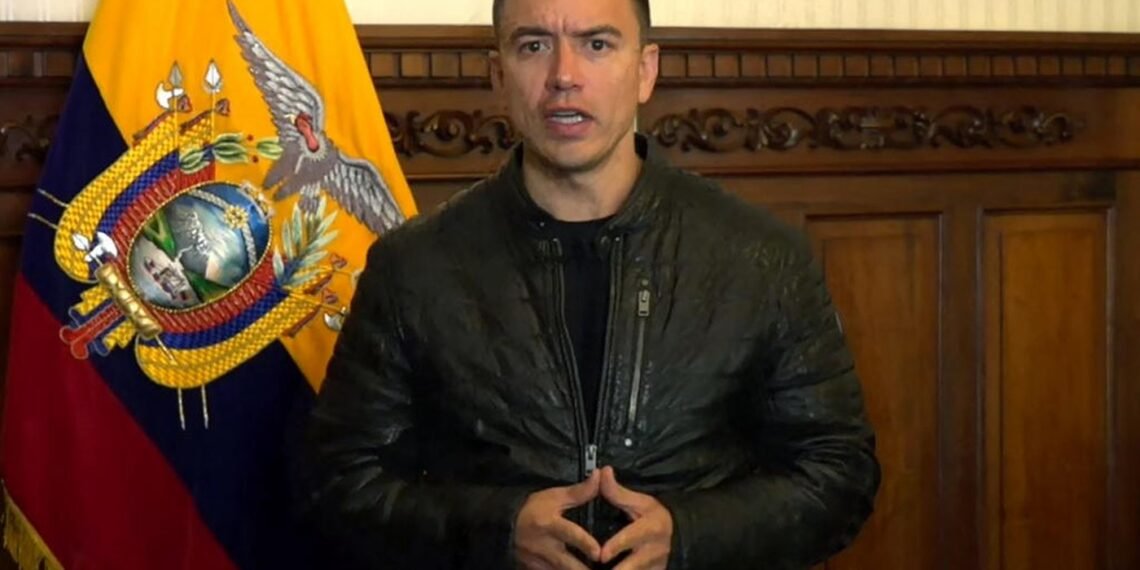 Homem em jaqueta preta fala seriamente em frente à bandeira do Equador, com madeira entalhada ao fundo.