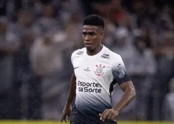 Jogador de futebol do Corinthians em campo, vestindo uniforme branco durante partida sob chuva intensa. Público ao fundo.