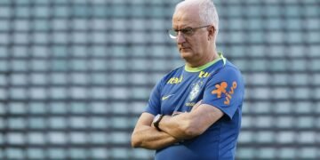 Treinador de futebol da seleção brasileira em campo, usando uniforme azul, olhando pensativo com os braços cruzados.