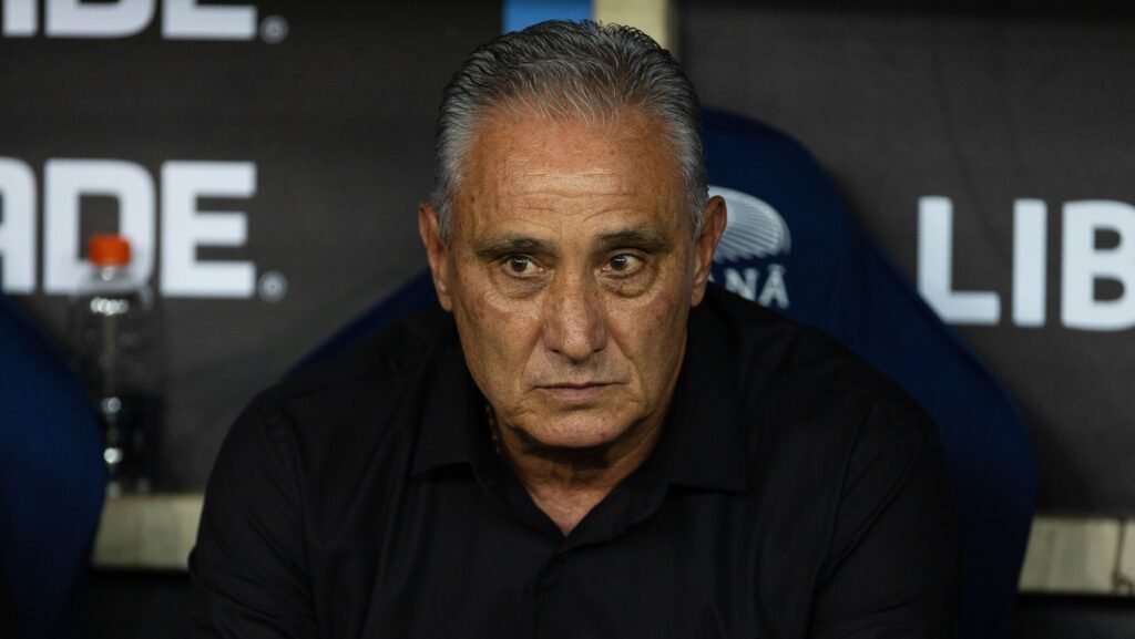 Homem de expressão séria em cadeira de estádio, ao fundo parede com logotipos parcialmente visíveis.