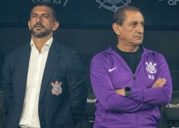 Dois homens em pé no banco de reservas do Corinthians, um com uniforme roxo e outro de terno com brasão do clube.
