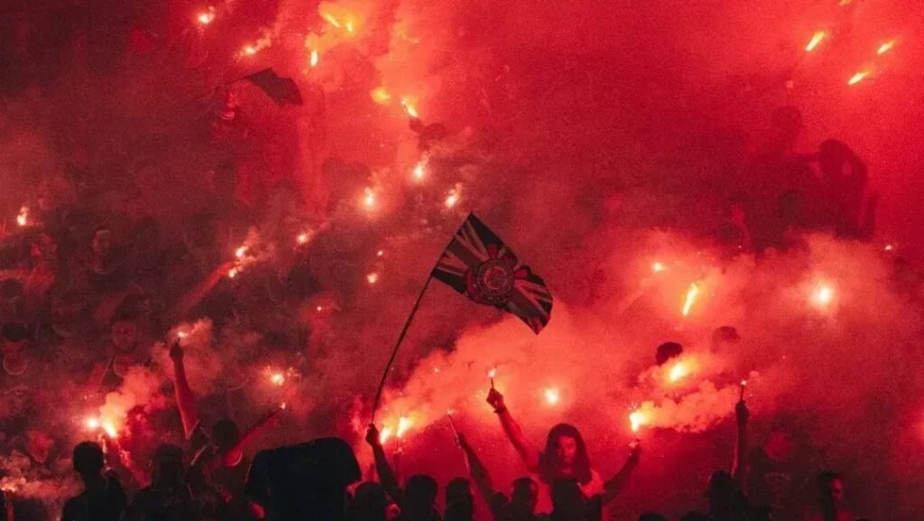 Torcida vibrante agita bandeiras e sinalizadores em estádio, atmosfera intensa e vermelha destaca paixão pelo futebol.
