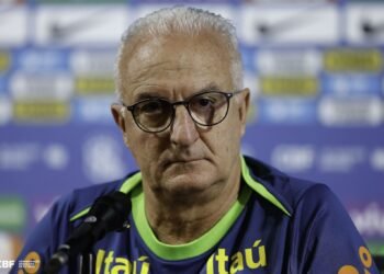 Homem de óculos em coletiva de imprensa, com fundo de logomarcas esportivas, aparenta concentração e seriedade.